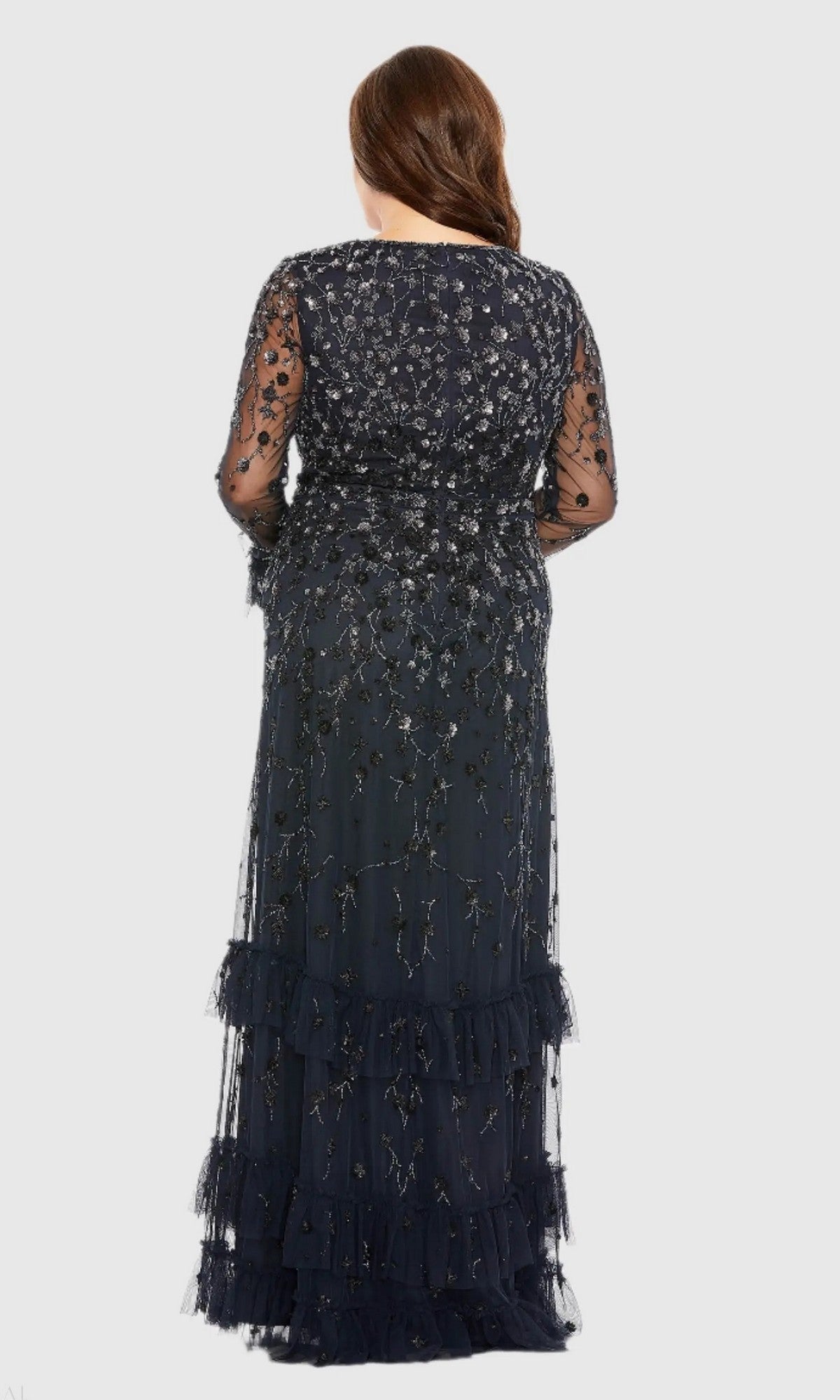 A woman stands facing away, wearing the Mac Duggal 9271 long plus-size formal dress in dark blue, featuring floor-length sequin beading, sheer beaded long sleeves, and intricate embroidery. Her hair is styled in loose waves down her back.